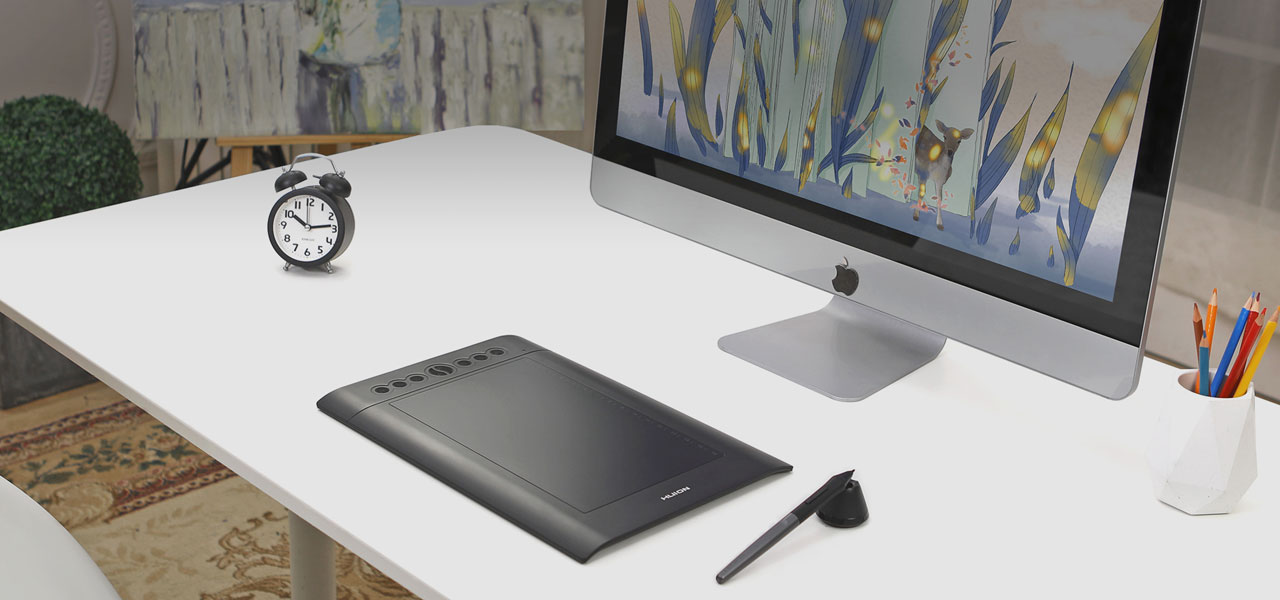 A Huion H610PRO V2 Drawing Tablet on display on a desk, in front of an iMac. Next to the drawing tablet on the desk are a pen leaning on a pen stand, a pen cup with multiple pencils in, and an alarm clock.